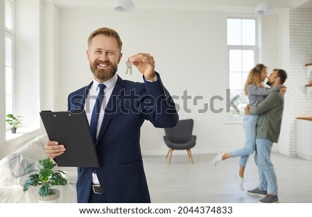 Realtor or real estate agent shows key to house he has sold. Portrait of man in suit standing in living room with happy married couple in background, holding folder and key, smiling, looking at camera Royalty-Free Stock Photo #2044374833