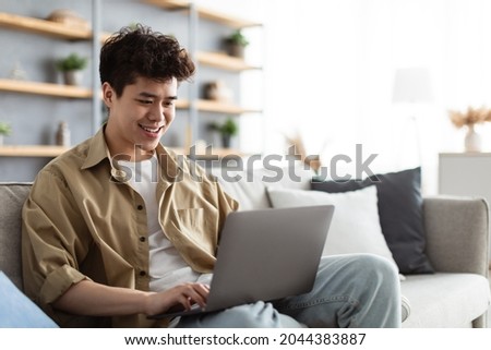 Portrait Of Smiling Asian Man Sitting On The Couch Working On Pc Laptop Indoors In Living Room. Happy Male Using Computer For Rest, Study Or Education, Typing On Keyboard, Surfing Internet Royalty-Free Stock Photo #2044383887