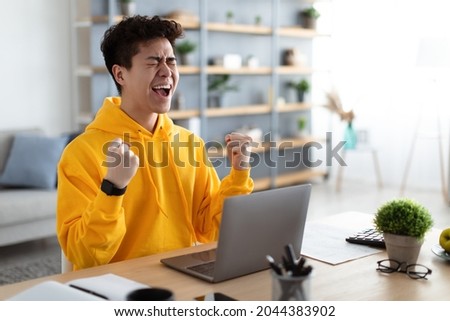 Excited asian man emotionally celebrating success using laptop in home office, sitting at desk raising hands with joy, shaking fists, received good news, got promotion, finished project, free space Royalty-Free Stock Photo #2044383902