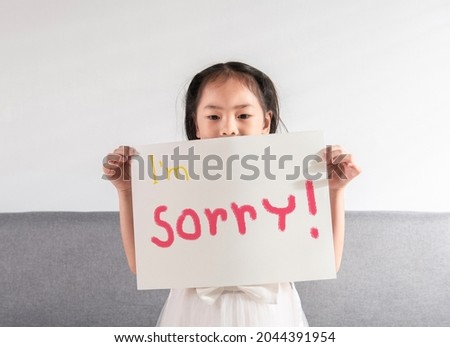 Cute Asian kid little toddler girl feels guilty is holding a sign of "I'm sorry" written on a white paper to apologize to her mother, father, or teacher for her mistake at home or school. Royalty-Free Stock Photo #2044391954