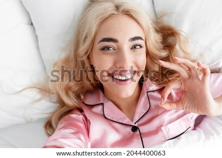 Cheerful millennial european blonde female in pink pajama shows ok gesture with hand, lies on bed and enjoys great morning, close up. Modern blogger and life stream, positive mood and facial emotions
