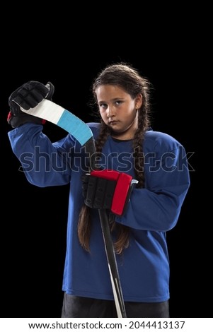 Portrait of one young girl professional hockey player in blue uniform, holding stick isolated over black background. Youth culture. Concepth of childhood, sport, strength ad