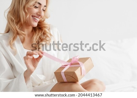 Glad young attractive european blonde female in robe, opens gift box, unties ribbon on white bed in bedroom, close up, copy space. Anniversary, happy birthday, Valentine day and positive emotions Royalty-Free Stock Photo #2044413467