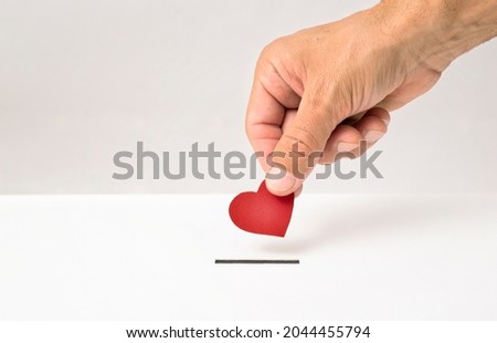 Red heart symbol is put by person's hand into slot of white donation box, Concept of donorship, life saving or charity Royalty-Free Stock Photo #2044455794