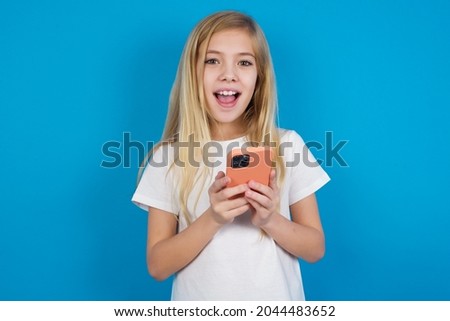 caucasian little girl wearing white T-shirt over blue background holding gadget while sticking out tongue