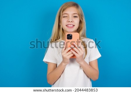 Pleased caucasian little girl wearing white T-shirt over blue background using self phone and looking and winking at the camera. Flirt and coquettish concept.