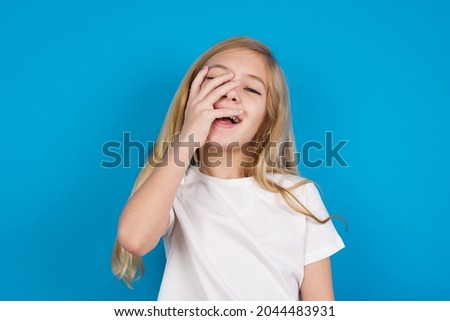 Charismatic carefree joyful caucasian little girl wearing white T-shirt over blue background likes laugh out loud not hiding emotions giggling hear funny hilarious joke chuckling facepalm.