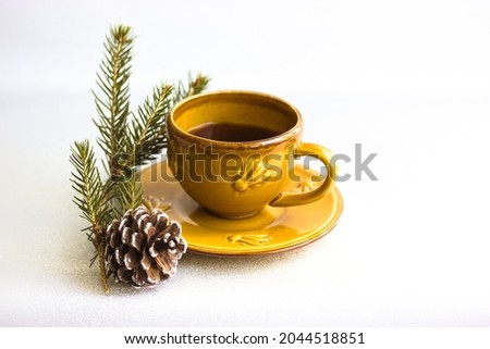 A small yellow ceramic coffee cup on a saucer stands on a white table background. Christmas tree branch and fir cone. Beautiful minimalist nature composition. Place for text. Cozy home. Winter hygge.
