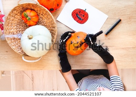 Preparing for holiday, decorating the house for Halloween. A child in black gloves draws a laughing face on a pumpkin, sitting at a desk, next to a pumpkins in a wicker basket and a sketch, top view.