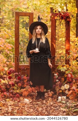 Young woman in Witch costume and hat looking mysteriously at candle in her hands isolated against autumn forest background with vintage wooden doors used as staged decoration for Halloween photo shoot