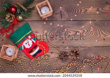 Red felt stocking with the image of Santa Claus, fir branches, cones and Christmas tree decorations, a candle, gold beads on a wooden table. Top view, copy space.