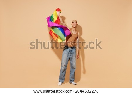 Full body young activist smiling happy fun blond latin gay man with make up in beige tank shirt waving hold rainbow flag isolated on plain light ocher background studio People lgbt lifestyle concept. Royalty-Free Stock Photo #2044532276
