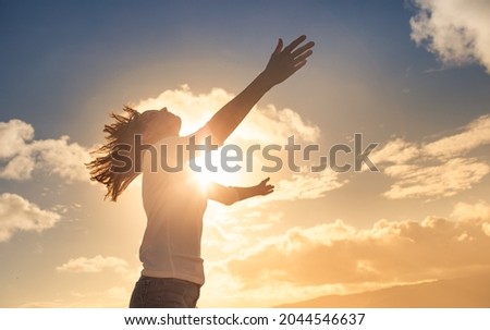 Young woman looking up feeling energized by the warm rays of sunshine lifting arms up to the sunset sky. Letting go of your fears concept.  Royalty-Free Stock Photo #2044546637