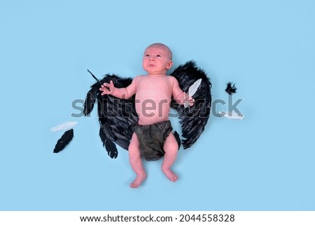 A newborn baby boy with dark evil wings on a blue studio background, copy space. An infant caucasian child in a diaper aged one month