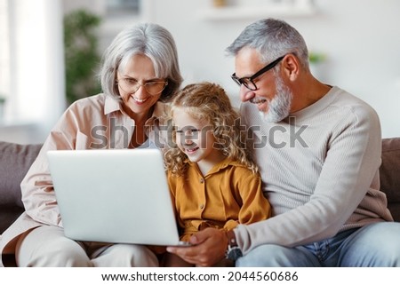 Smiling senior grandparents and cute little girl granddaughter looking at laptop computer screen while sitting on sofa at home, happy child watching cartoons with grandpa and grandma on weekend