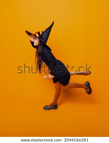 Little girl dressed as a Halloween witch in a black dress and hat flies on a broomstick on a yellow background.