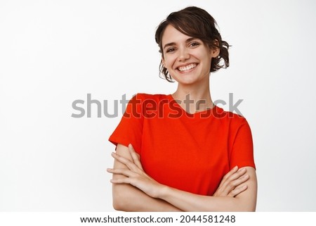 Beautiful stylish woman cross arms on chest, looking confident and smiling at camera, standing in red t-shirt against white background