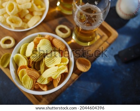 Light beer in glasses, potato chips, onion rings on a wooden tray, a baseball and a TV remote. High angle view. Watching sports games, matches, TV programs with friends, rest, relaxation.