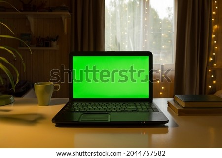 Laptop Computer with Green Mock-up Screen. Green Screen Device Stands on a Desk in the Living Room. In the Background Cozy Home with Christmas Decoration in the Evening with Warm Lights on