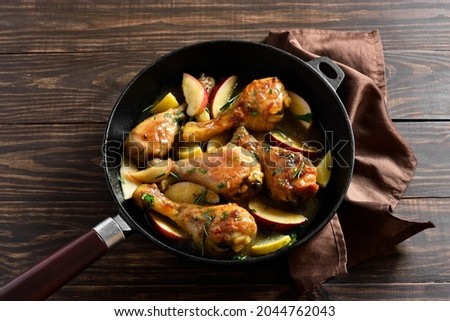 Close up view of chicken drumsticks baked with apples and herbs in frying pan on wooden table.