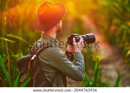 Backpacker man traveler photographer holding a dslr camera in hands standing in field at sunset 