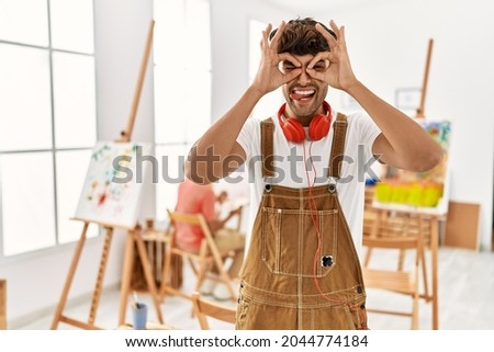 Young hispanic man at art studio doing ok gesture like binoculars sticking tongue out, eyes looking through fingers. crazy expression. 