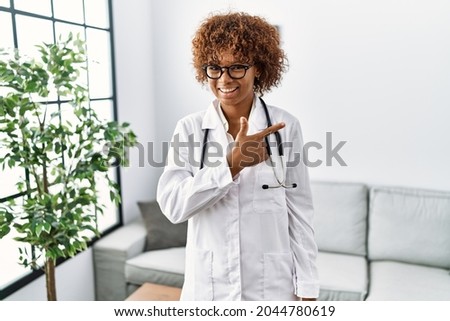 Young african american woman wearing doctor uniform and stethoscope cheerful with a smile of face pointing with hand and finger up to the side with happy and natural expression on face 