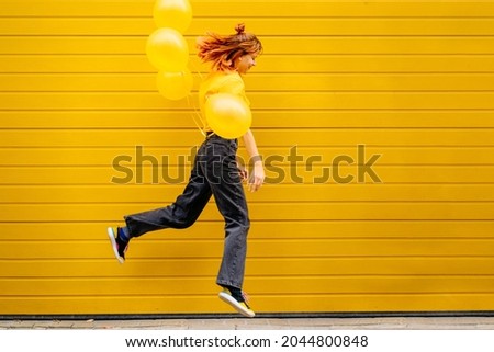 Full height of adult hipster girl having fun with balloons, jumping and flying with helium yellow balloons on yellow wall on background in the city street outdoor.