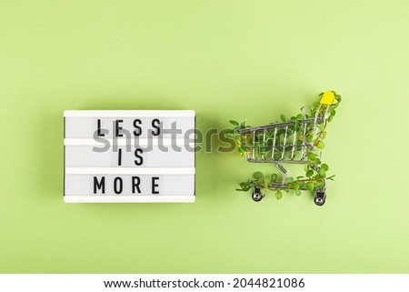 Less is more - written on lightbox next to Shopping cart entwined with plants on green background. Conscious consumption slow fashion Zero waste concept. Top view flat lay Royalty-Free Stock Photo #2044821086
