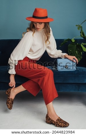 Autumn fashion conception: elegant woman wearing stylish orange hat, wide culottes, white silk blouse, leopard print loafer shoes, holding blue faux leather bag Royalty-Free Stock Photo #2044823984