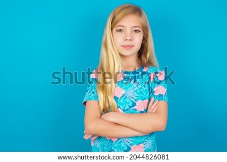 Confident Caucasian blonde kid girl wearing hawaiian T-shirt against blue wall with arms crossed looking to the camera