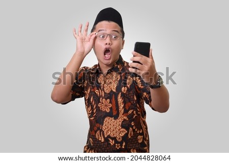 Portrait of shocked Asian man wearing batik shirt and songkok looking at his mobile phone. Wow face expression. Isolated image on gray background