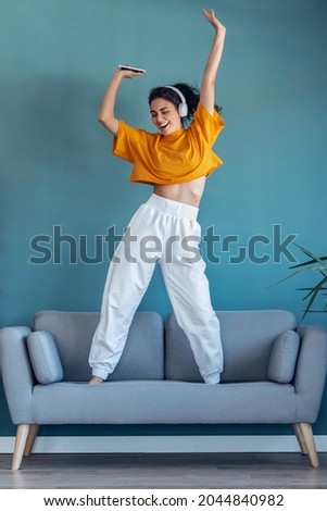 Shot of funny woman listening to music with smartphone while dancing on the couch in the living room at home.