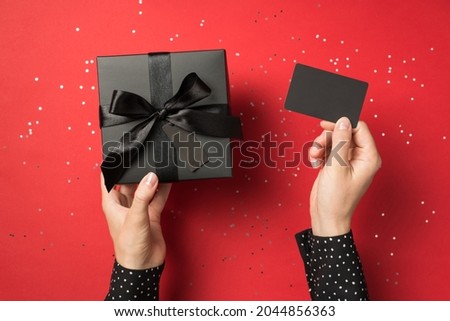 First person view overhead photo of black giftbox with ribbon wrapped as bow and business card in hands and confetti around isolated on the red background