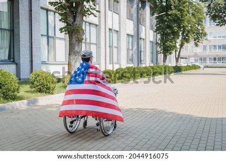 back view of disabled soldier, wrapped in usa flag, sitting in wheelchair on urban street
