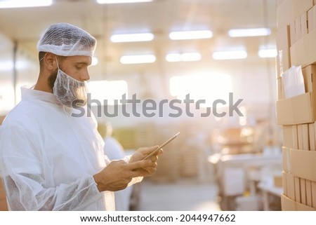A supervisor with a hairnet and beard net in a sterile white uniform is standing next to a pile of boxes and checking them. He holds the tablet in hand and looking at it. Quality control Royalty-Free Stock Photo #2044947662