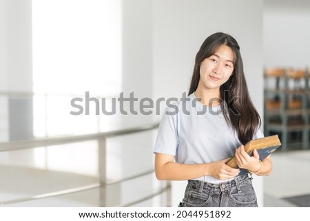 Front view portrait of a young cheerful Asian adolescent woman college student in relaxed casual back to school carry books stands in campus building