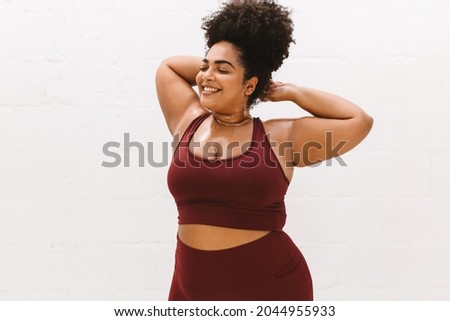Portrait of healthy woman with sweat on body and smiling with closed eyes while relaxing after workout. Fitness female in exercise outfit standing against white background. Royalty-Free Stock Photo #2044955933