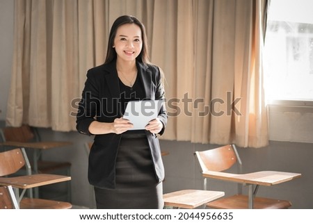 Adult confident cheerful Asian woman teacher in black business uniform using digital tablet and laptop sitting in classroom