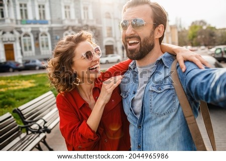 attractive laughing man and woman traveling together, stylish couple in love taking selfie photos on phone on romantic trip, sunny summer city, wearing shirt, sunglasses, travelers having fun