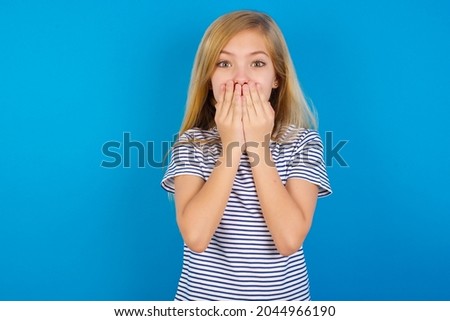 Vivacious Caucasian blonde kid girl wearing stripped T-shirt against blue wall, giggles joyfully, covers mouth, has natural laughter, hears positive story or funny anecdote