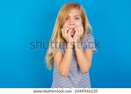 Fearful Caucasian blonde kid girl wearing stripped T-shirt against blue wall keeps hands near mouth, feels frightened and scared,  has a phobia,  Shock and frighted concept.