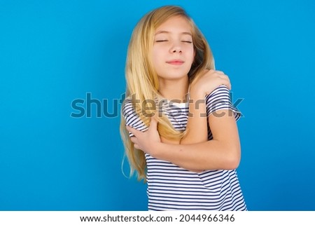 Caucasian blonde kid girl wearing stripped T-shirt against blue wall Hugging oneself happy and positive, smiling confident. Self love and self care