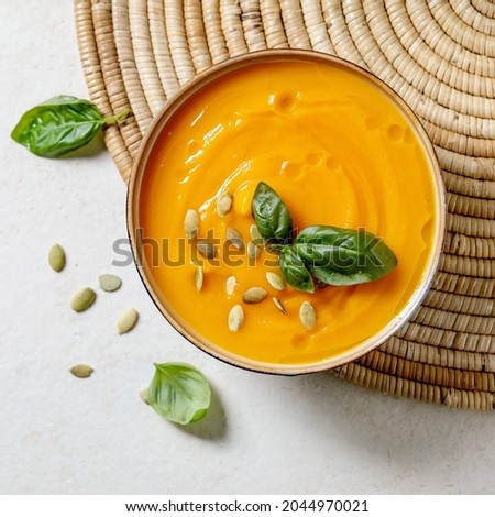 Bowl of pumpkin or carrot vegetarian cream soup decorated by fresh basil, olive oil and pumpkin seeds on white texture background with ingredients above. Flat lay, copy space. Square image