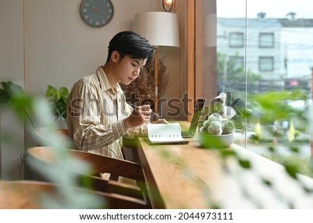Photo of a young smart businessman taking notes while sitting at the wooden counter surrounded by a laptop computer and various stuff.