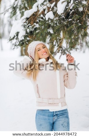 Beautiful girl in a Winter Suede Lambs Wool Jacket enjoying winter moments. Outdoors photo of blonde in a pink hat having fun on a snowy morning on a blurred nature background.