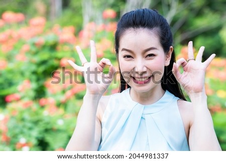 Beautiful charming asian young woman smiling and showing OK sign outdoors with natural of flower. Happy expression of lady hand make a sign OK. Woman has pleasant smile and good mood, Body language.