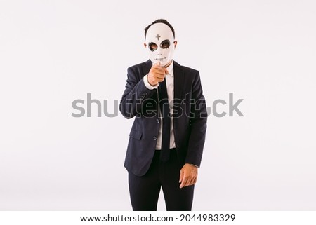 Man dressed in suit and tie, wearing a killer's mask with a cross on his forehead for Halloween, pointing with his finger. Carnival and halloween celebration