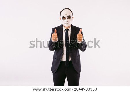 Man dressed in suit and tie, wearing a Halloween cross-forehead killer mask, thumbs up. Carnival and halloween celebration