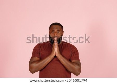Hopeful young African-American person actor with beard in brown t-shirt prays looking forward on pink background in studio closeup